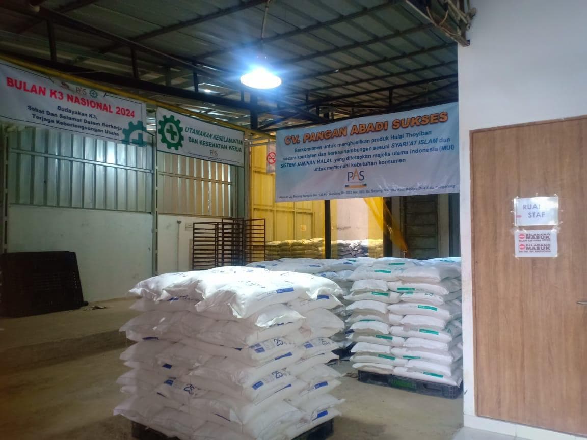 Tumpukan karung putih di gudang dengan spanduk keselamatan kerja dan pintu bertanda ruang staf.