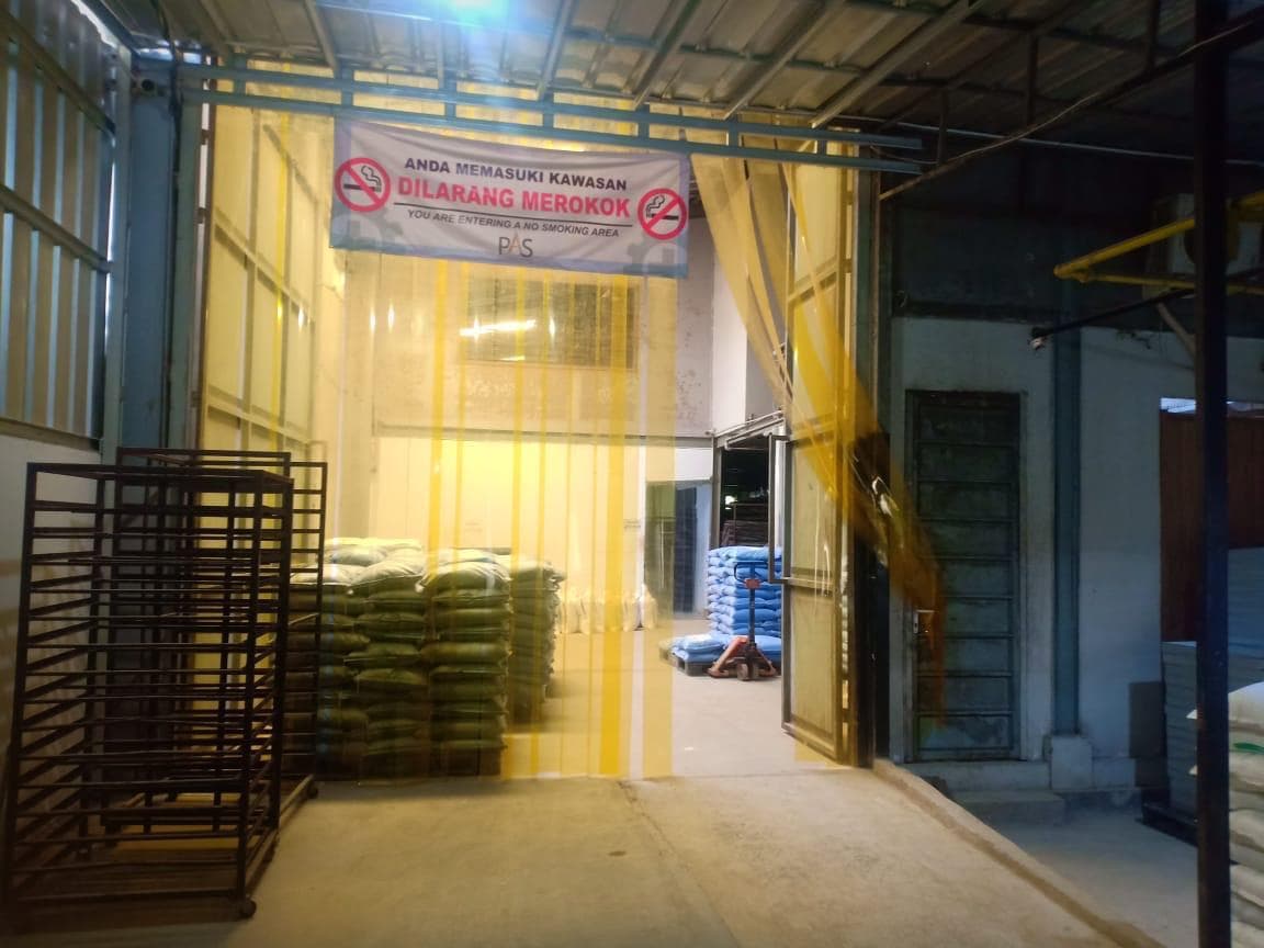Pintu masuk gudang industri dengan tirai plastik kuning, tumpukan karung, dan spanduk dilarang merokok.