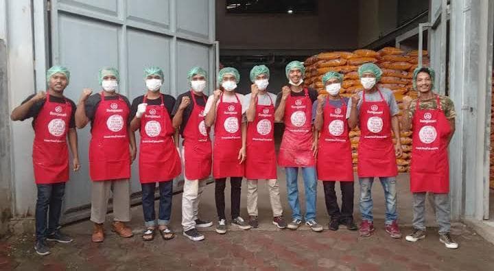 Sepuluh pekerja bercelemek merah dan penutup kepala berpose di depan tumpukan karung tepung.